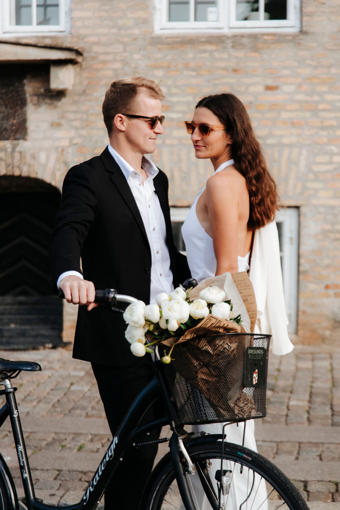 Stylish couple with white roses in bicycle basket posing against brick building.