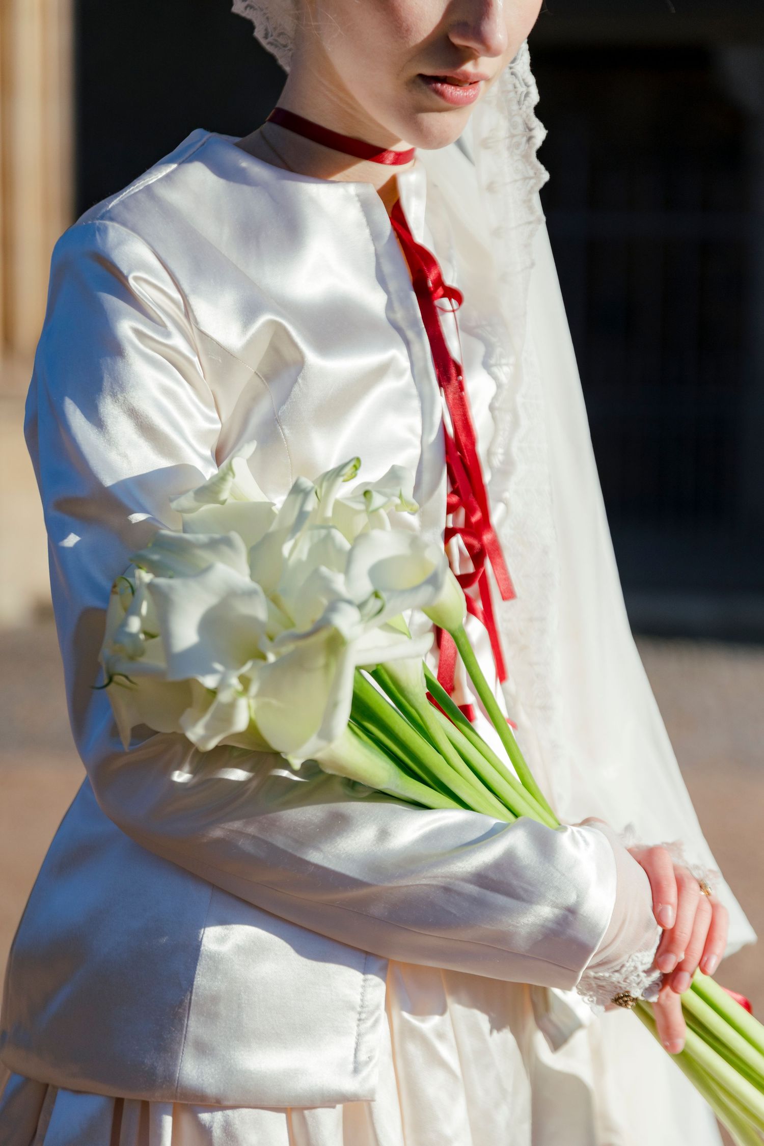 A person in a white dress holds elegant white calla lily bouquet with red ribbons.