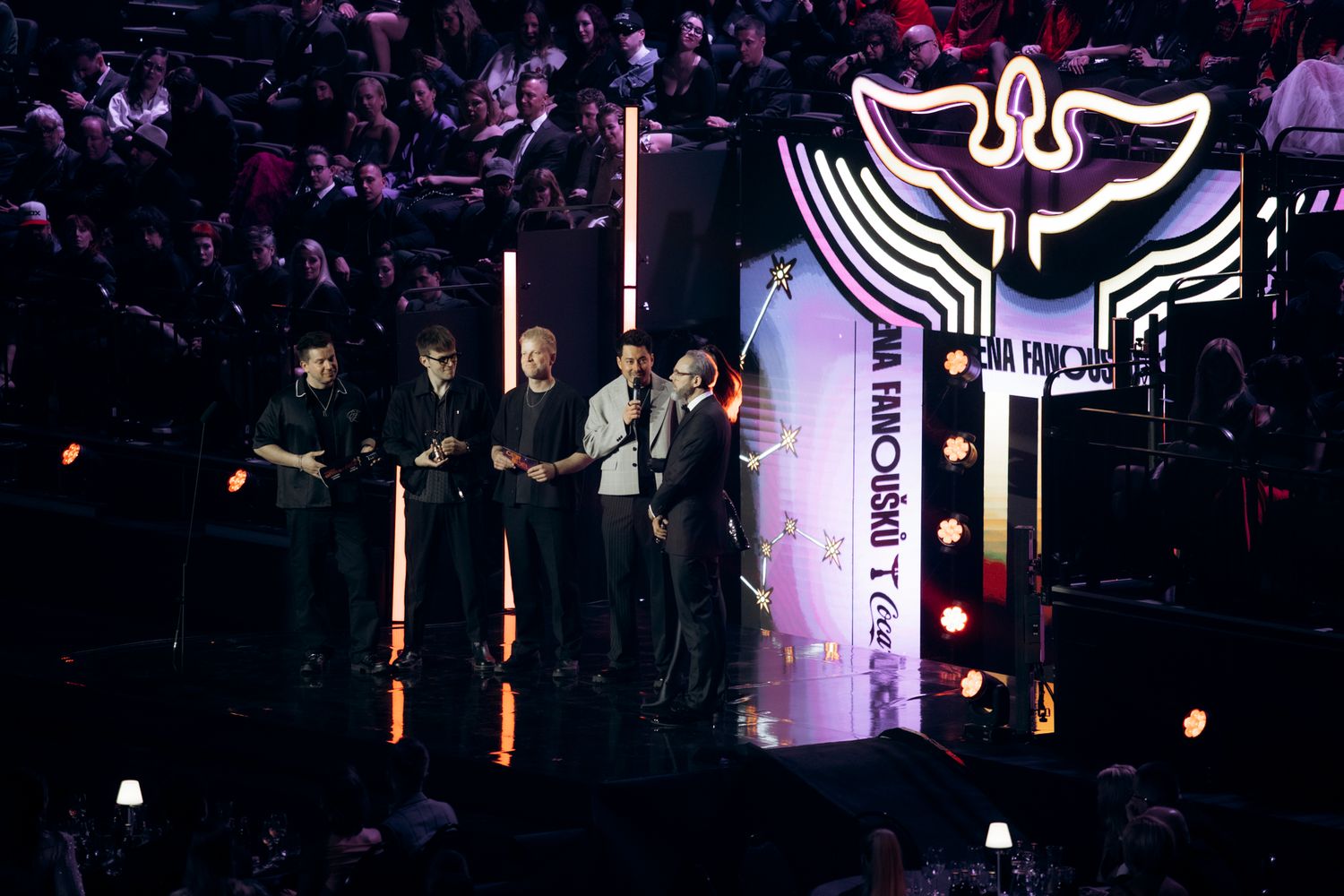 A dark concert venue illuminated with purple lighting and silhouettes of performers on stage during a live performance.