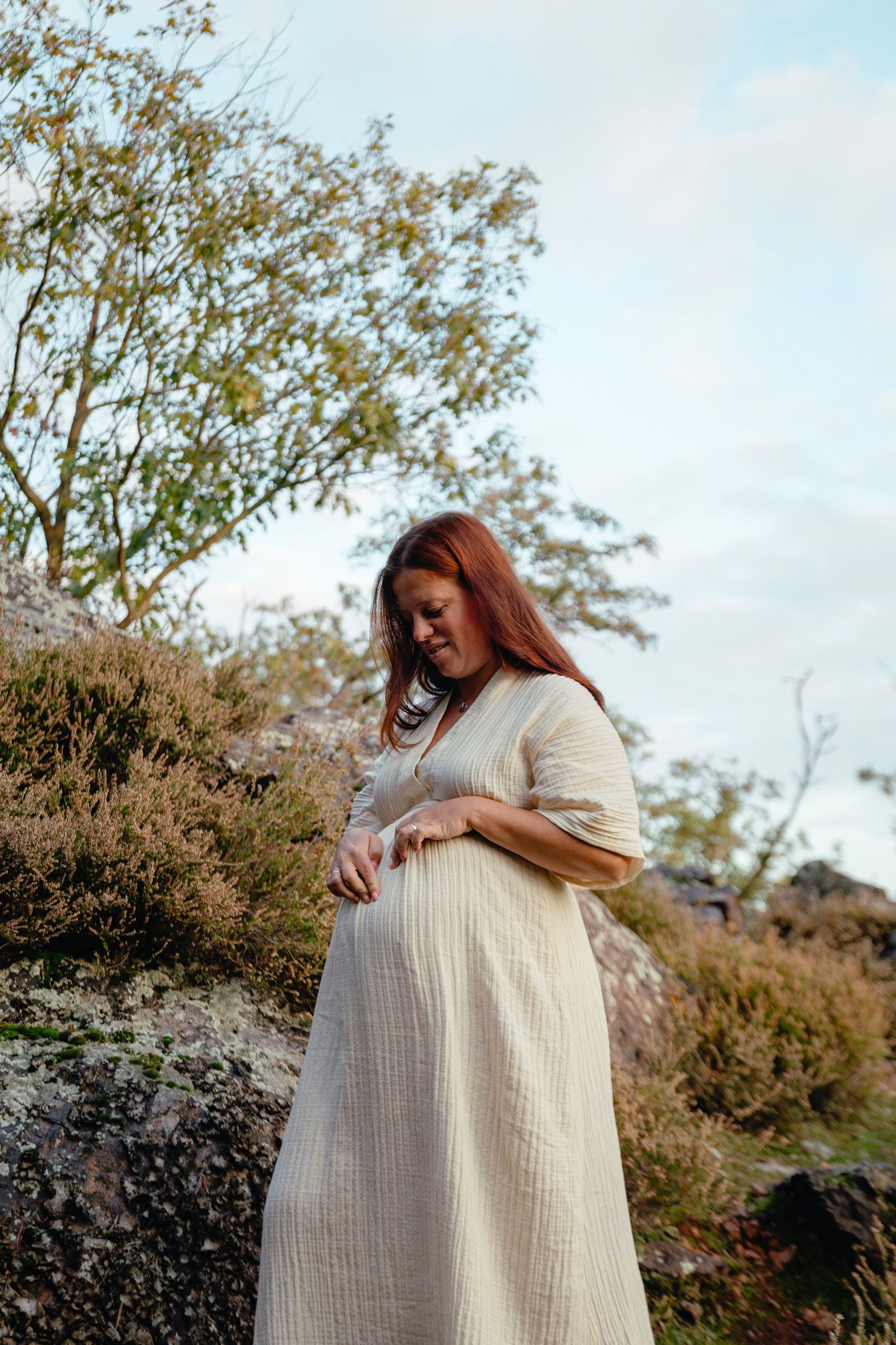 Woman in white dress cradling baby bump on rocky hillside at sunset.