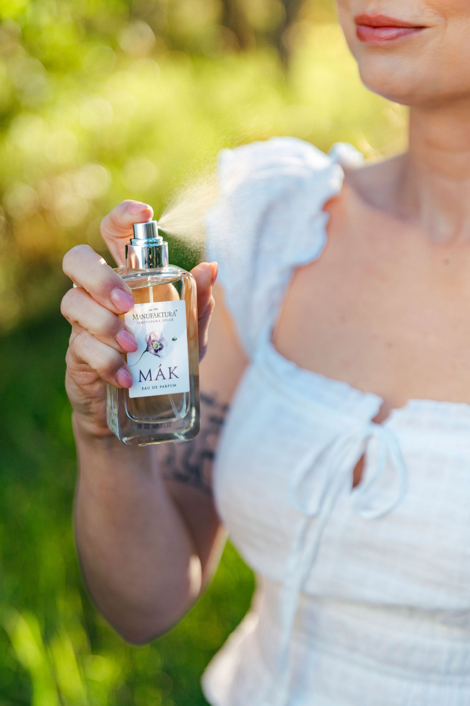 Woman in white dress holding a MAK perfume bottle by Hagmore outdoors in a green garden setting.