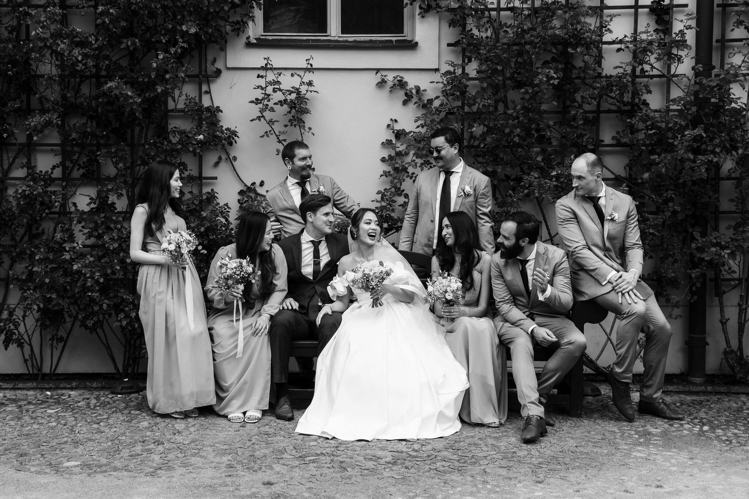 Wedding party relaxes on lawn outside historic building in black and white photo.