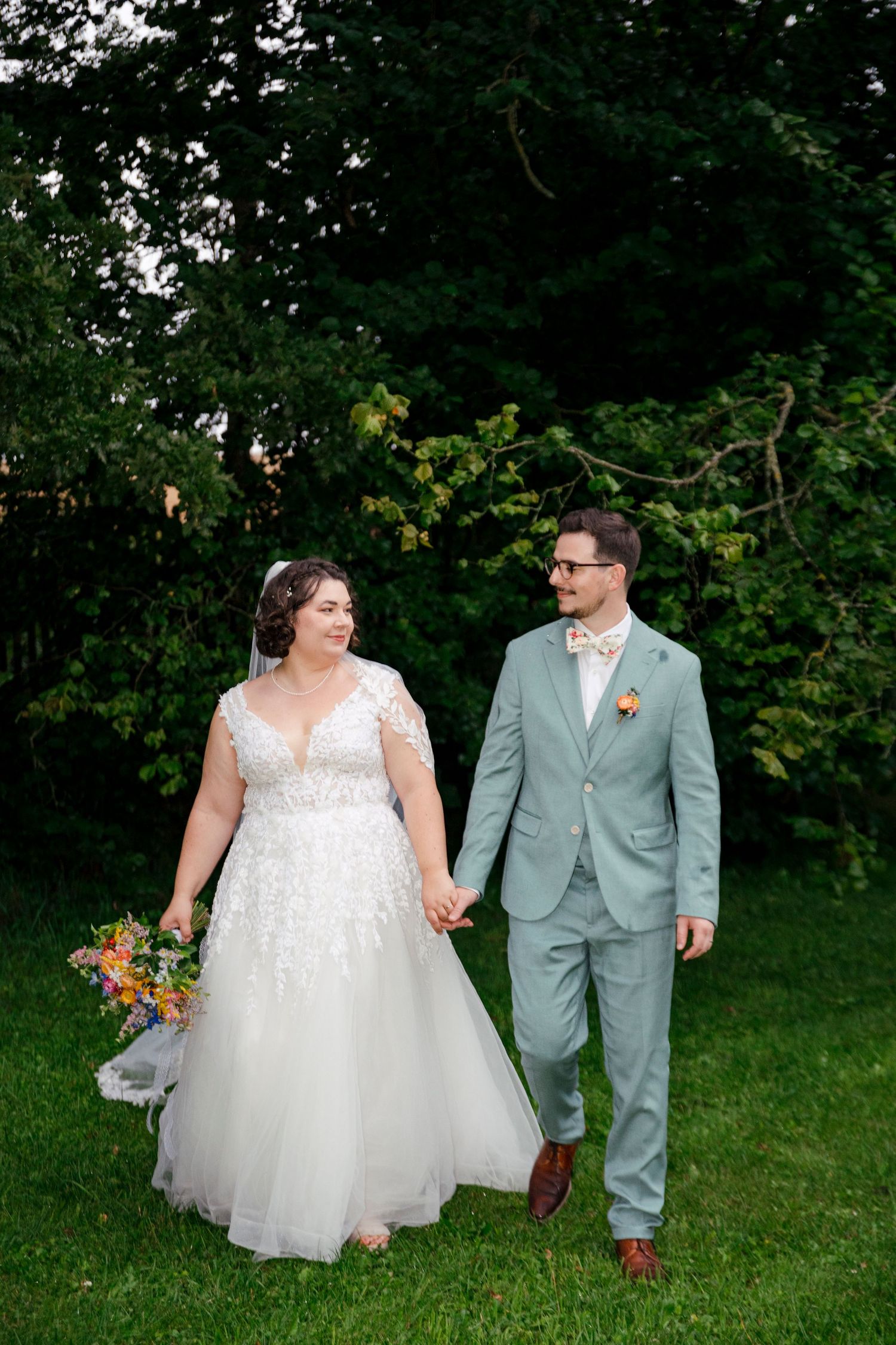 Couple walking together through a lush green garden, bride in white dress and groom in grey suit.