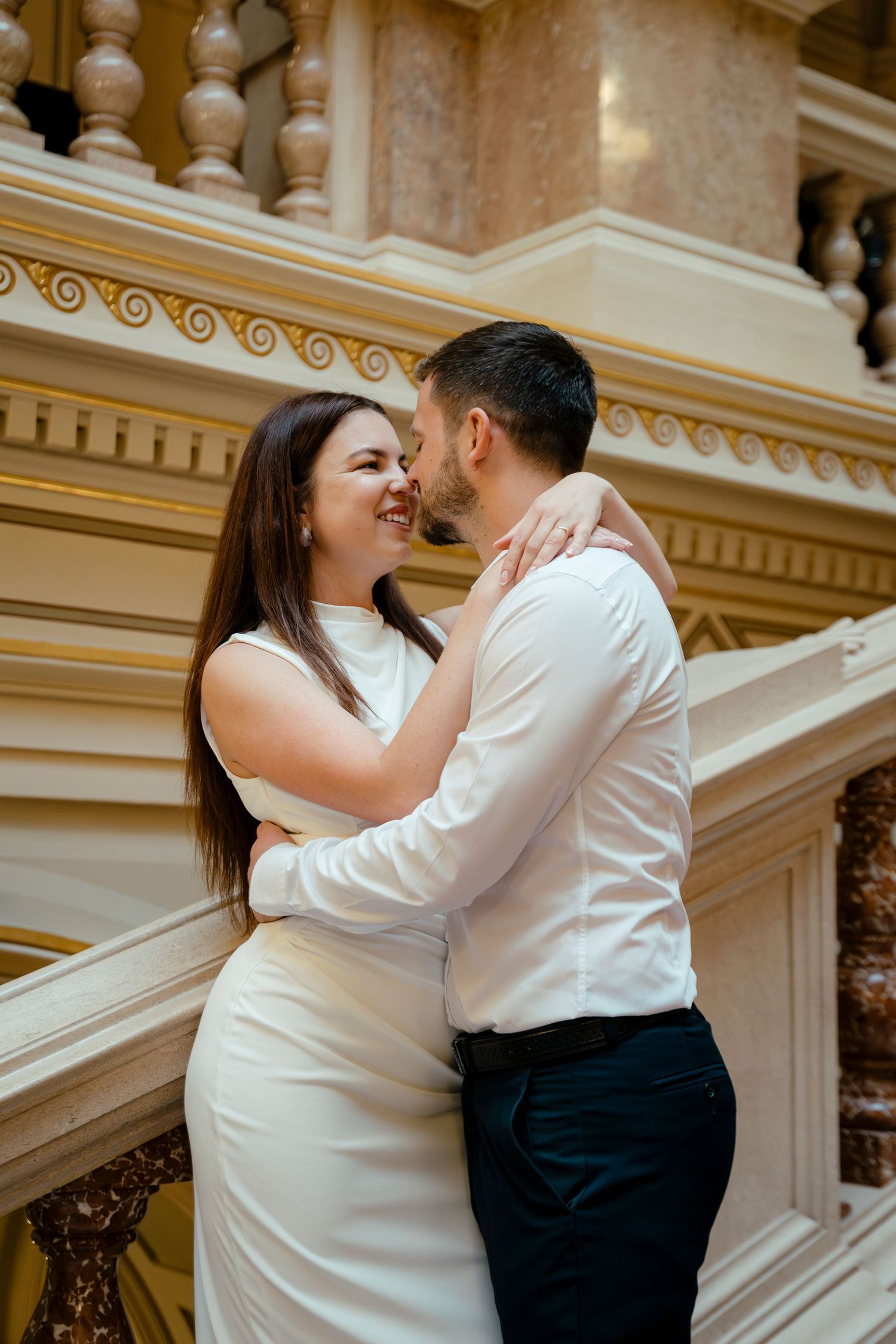 Couple embraces on ornate marble staircase in elegant historic building interior.