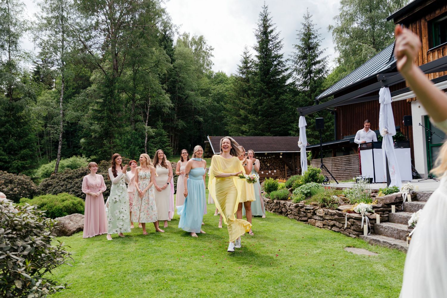 Bridesmaids in pastel dresses gather on green lawn outside rustic lodge for outdoor wedding bouquet toss.