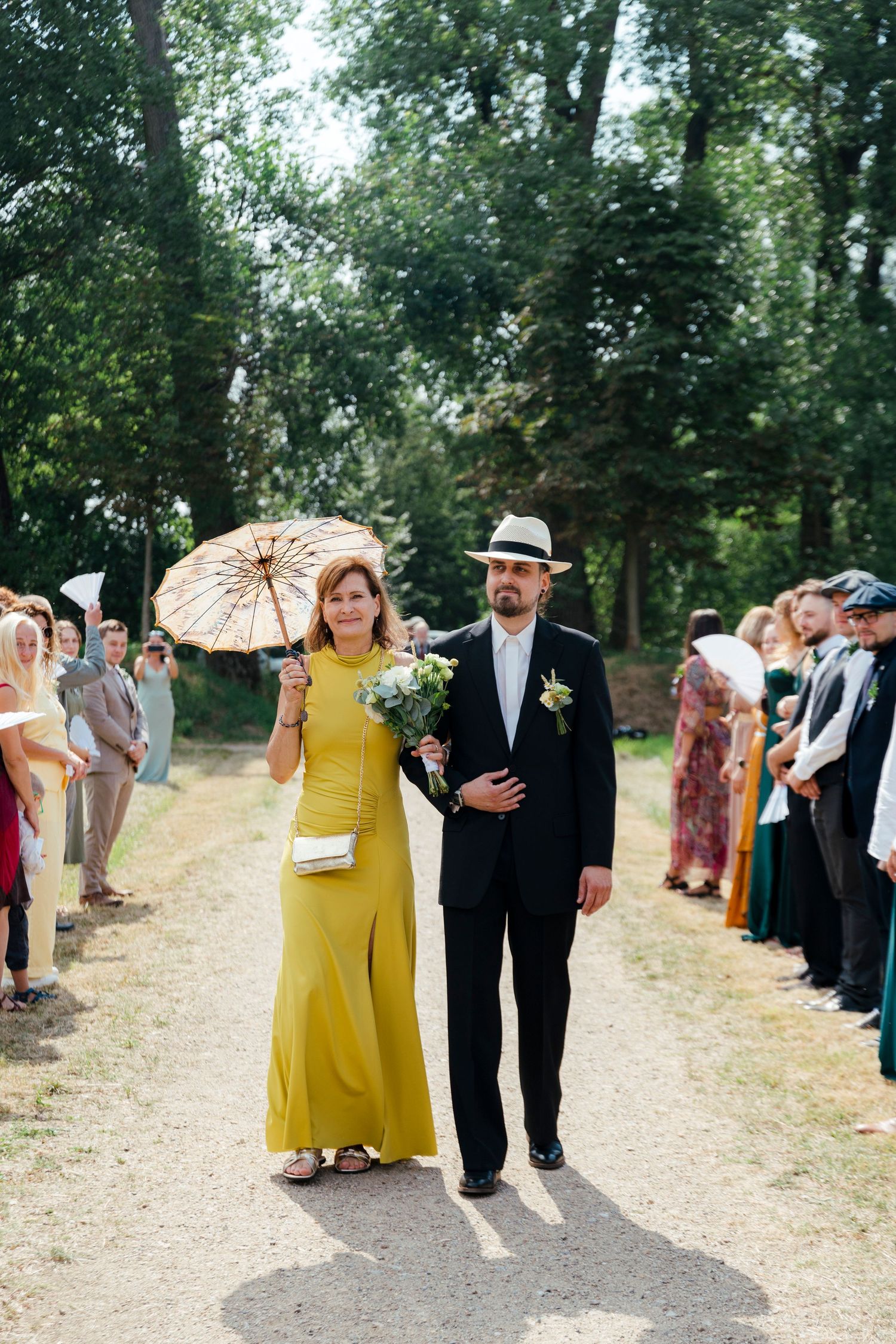 Elegant couple walking down outdoor wedding aisle, woman in yellow dress holding parasol and bouquet, man in black suit and hat.