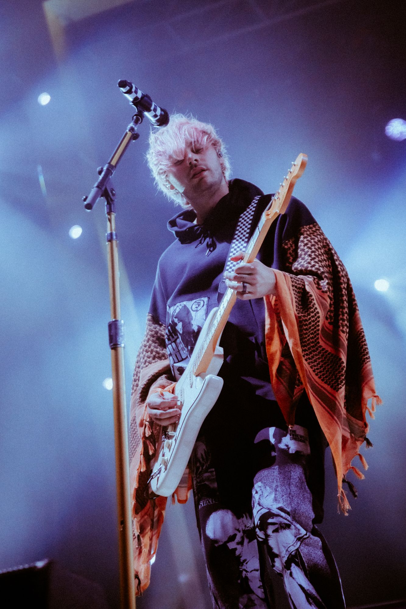 Guitarist performs on stage in dramatic lighting wearing a studded leather jacket and playing an electric guitar.