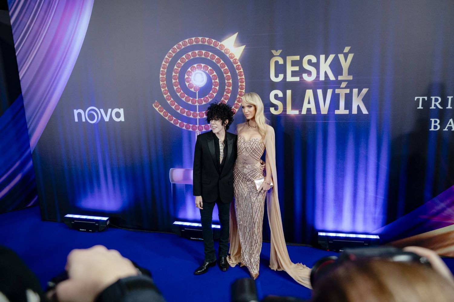 Fashion show models walk down a blue-lit runway displaying elegant evening gowns and formal attire.