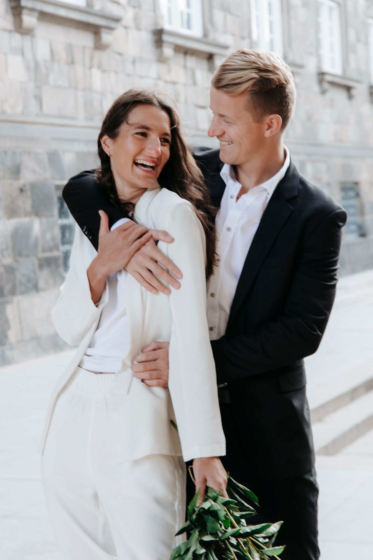 Couple sharing a joyful moment against stone wall in elegant wedding attire.