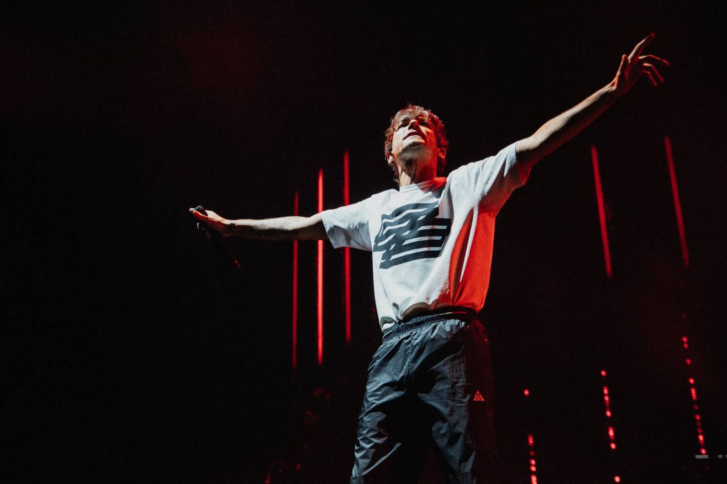Performer in white t-shirt and jeans strikes dramatic pose on dimly lit stage with red lighting effects.