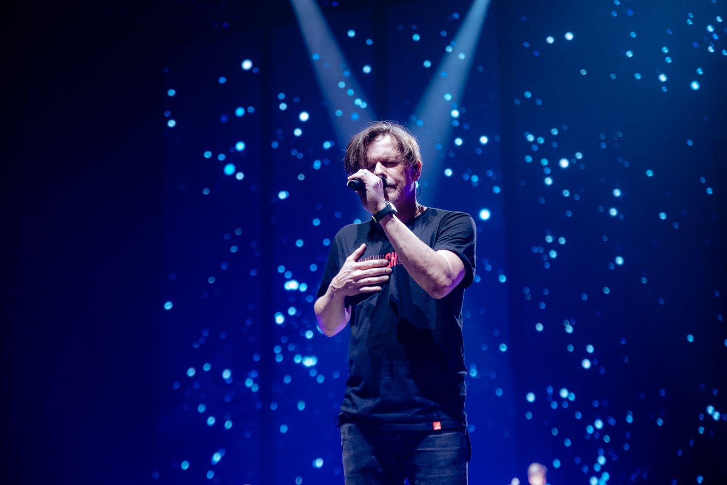Performer singing passionately on stage with dramatic blue lighting and sparkles in the background.