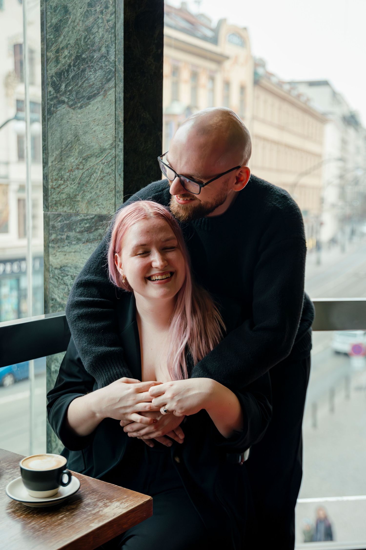 Couple sharing affectionate moment in cozy cafe with city views through window.