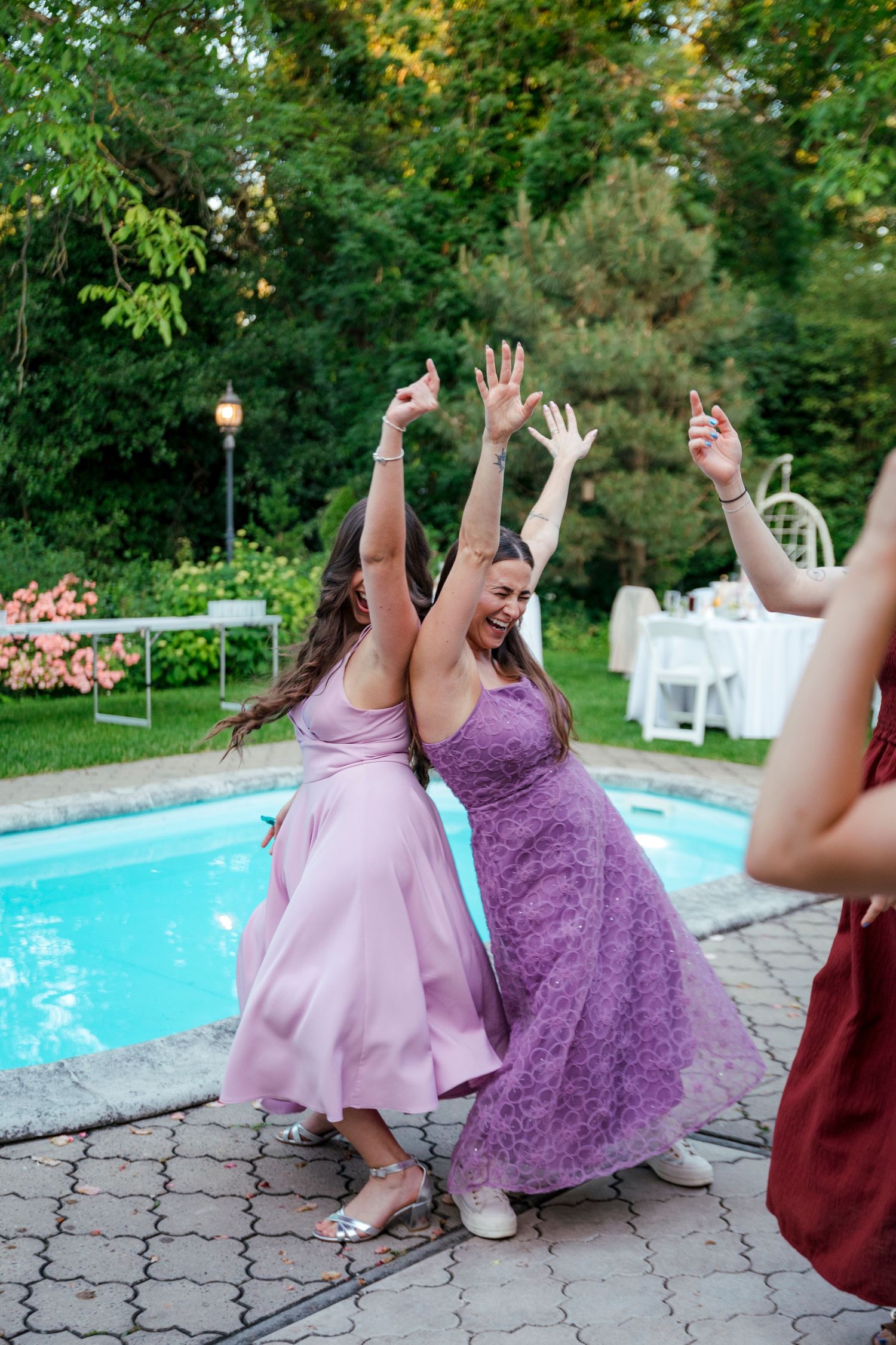 Bridesmaids in purple and pink dresses dancing joyfully at an outdoor wedding reception by a pool.