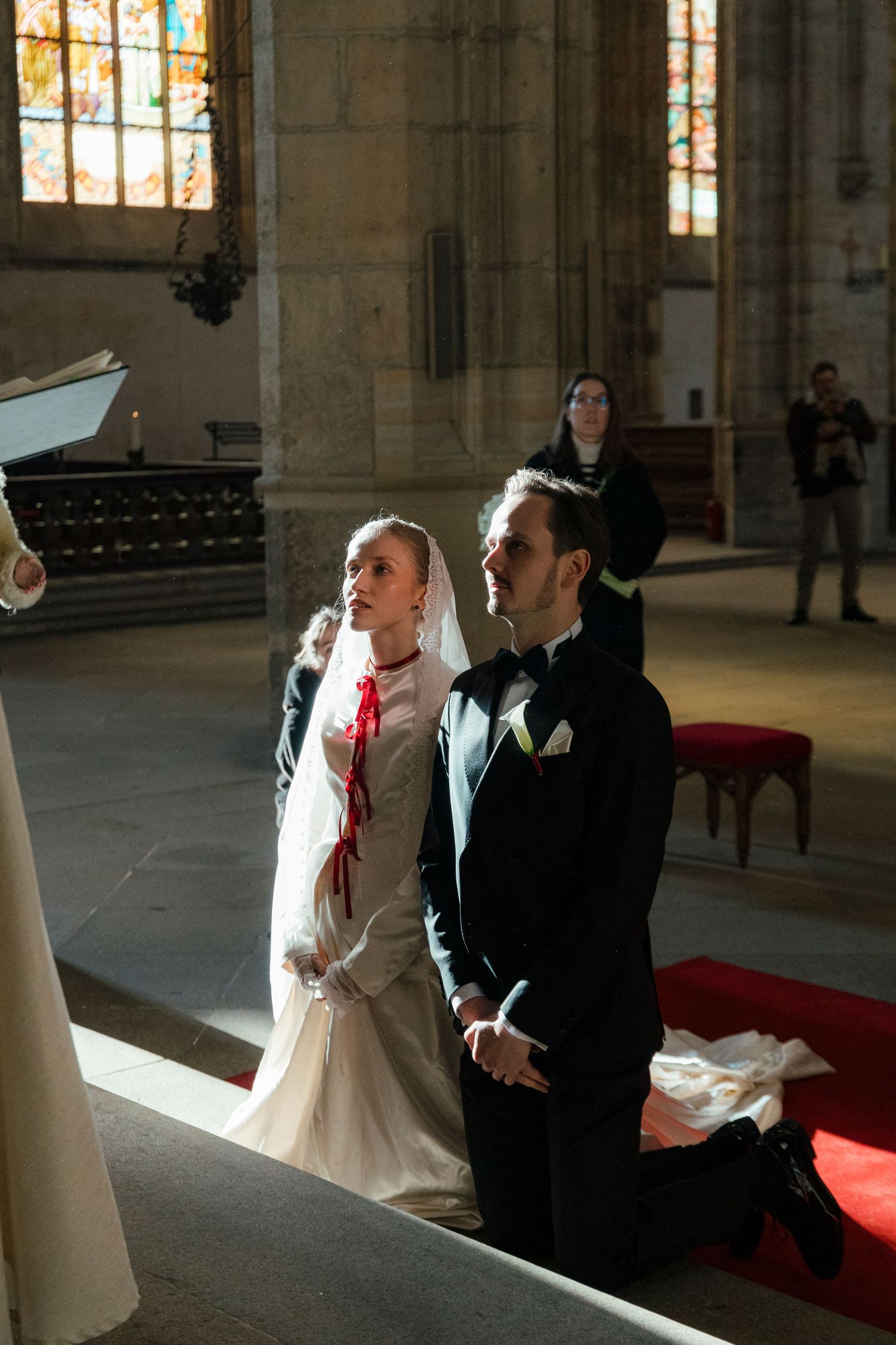 A couple in formal wedding attire share a tender moment inside a grand cathedral with stained glass windows.