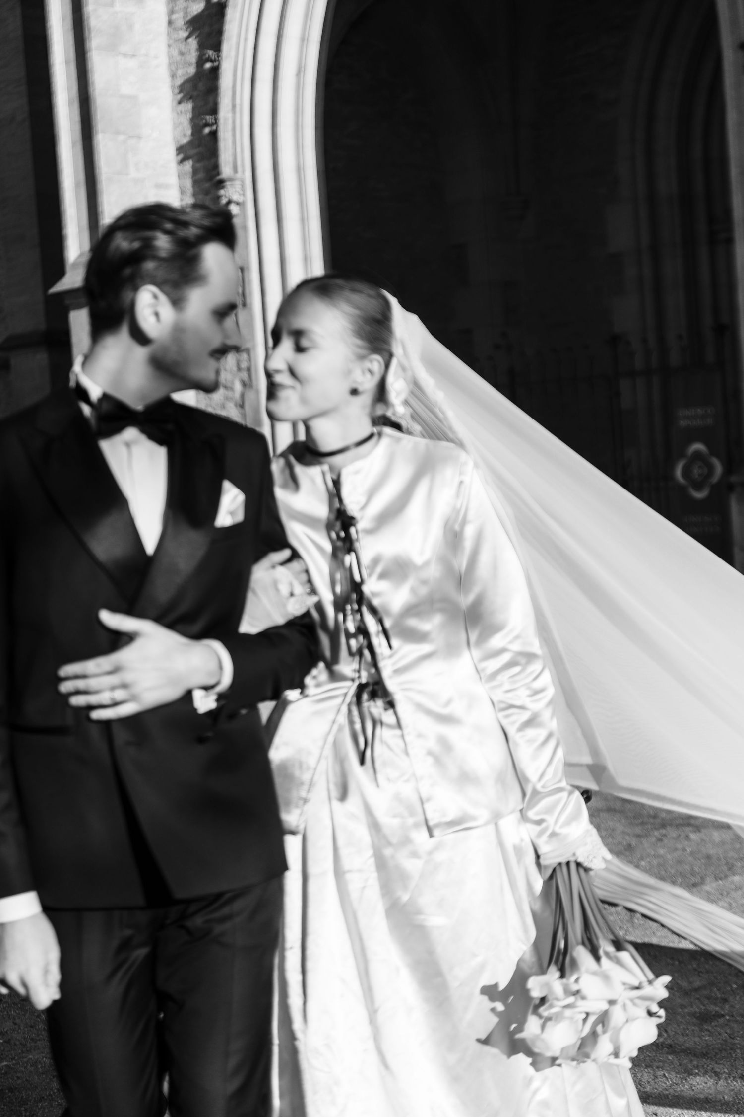 Black and white photo of a bride and groom sharing a romantic moment outside a church, bride in satin gown with veil.