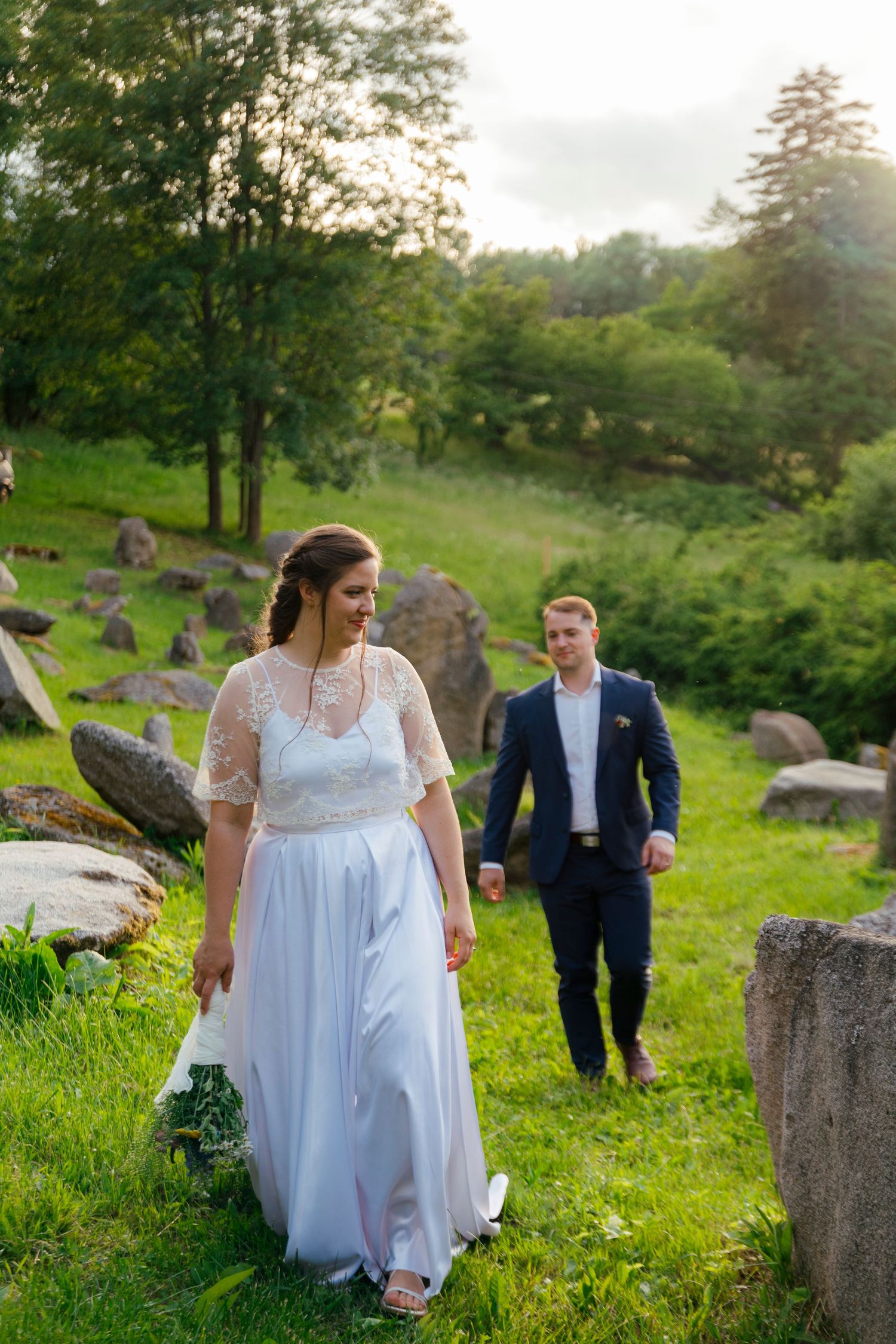 Bride and groom walking hand in hand on lush green hillside with scattered rocks during outdoor wedding photoshoot.
