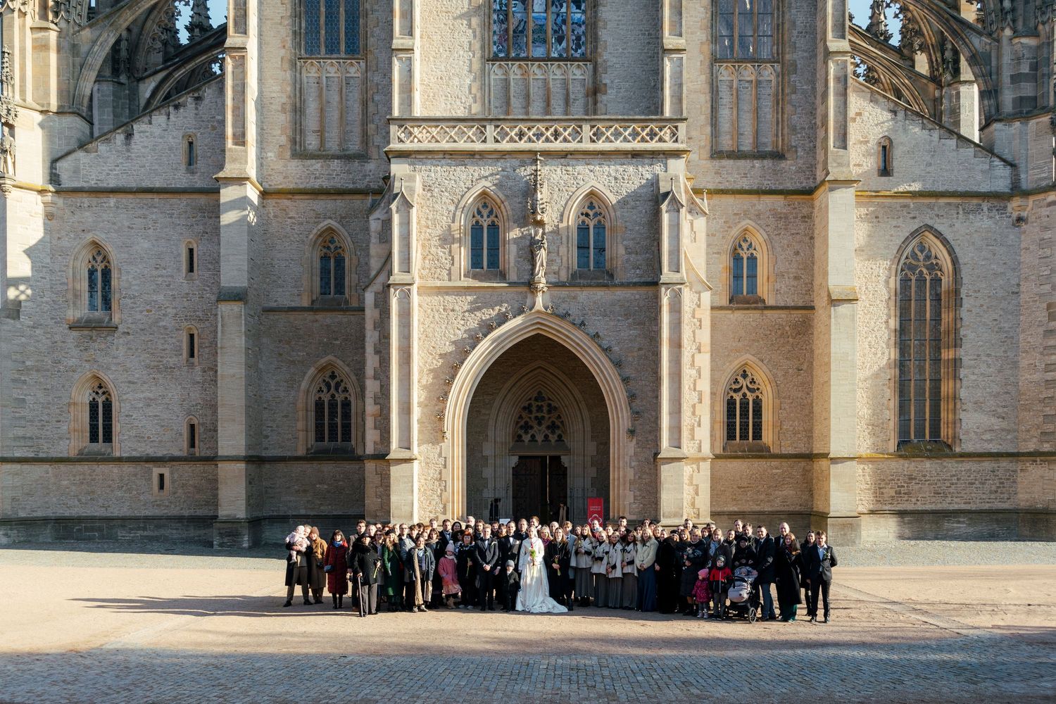 A panoramic view of a Gothic cathedral exterior with ornate architecture and groups of people gathered outside.