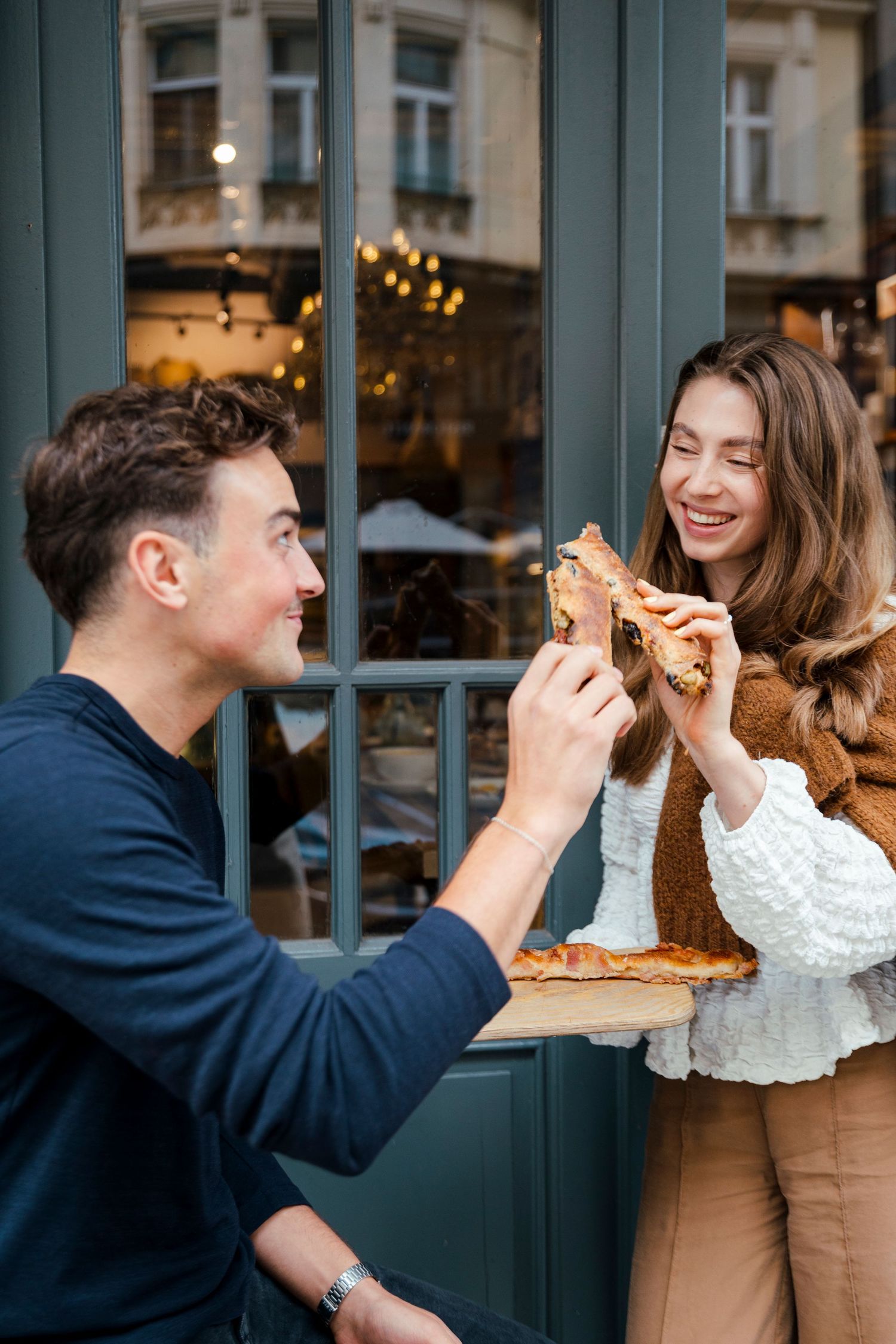 Two people sharing food and laughing outside a cafe storefront.