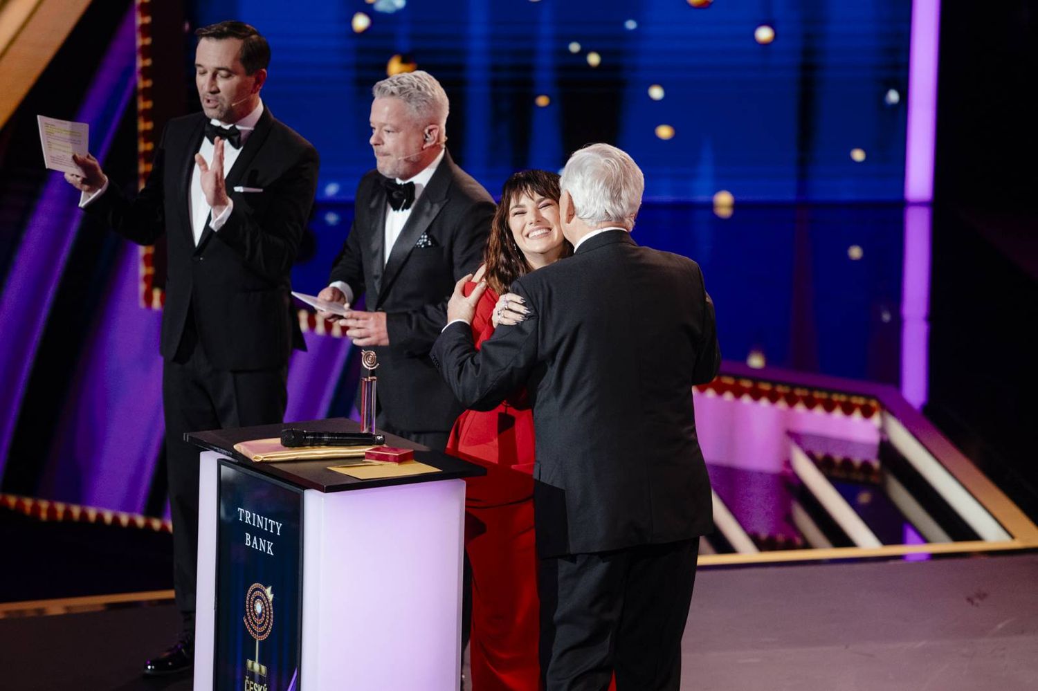 People in formal attire gather on stage during an awards ceremony with purple lighting and elegant decor.