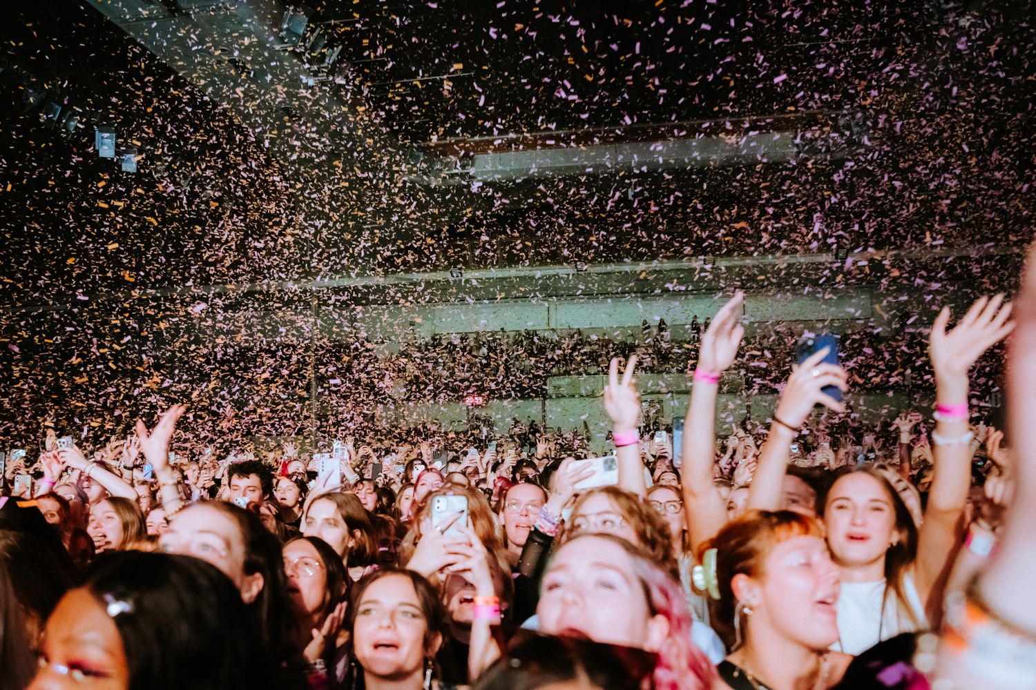 Concert crowd cheers with raised arms while colorful confetti rains down during a live music performance.