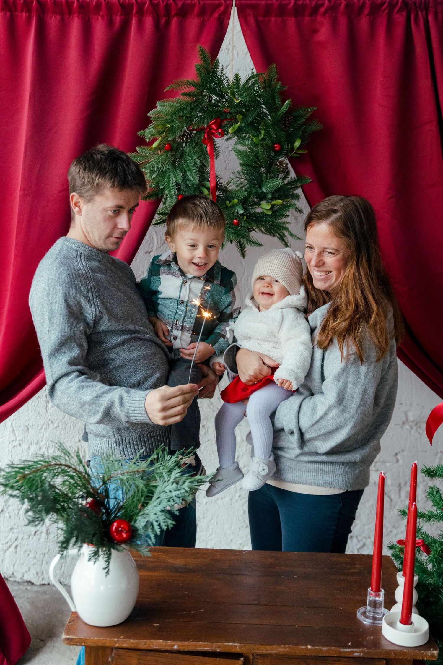 Happy family with two children celebrating Christmas, holding sparklers near festive wreath and red curtains.