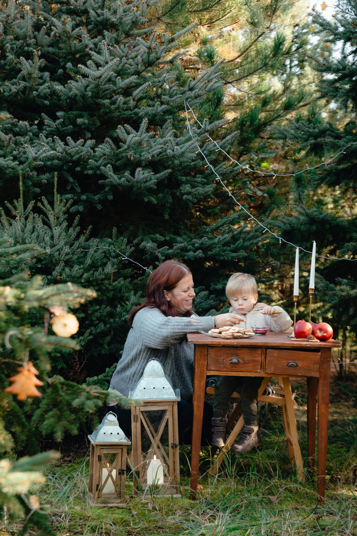 Mother and toddler sharing cookies at rustic outdoor table surrounded by evergreen trees with festive lanterns and candles.