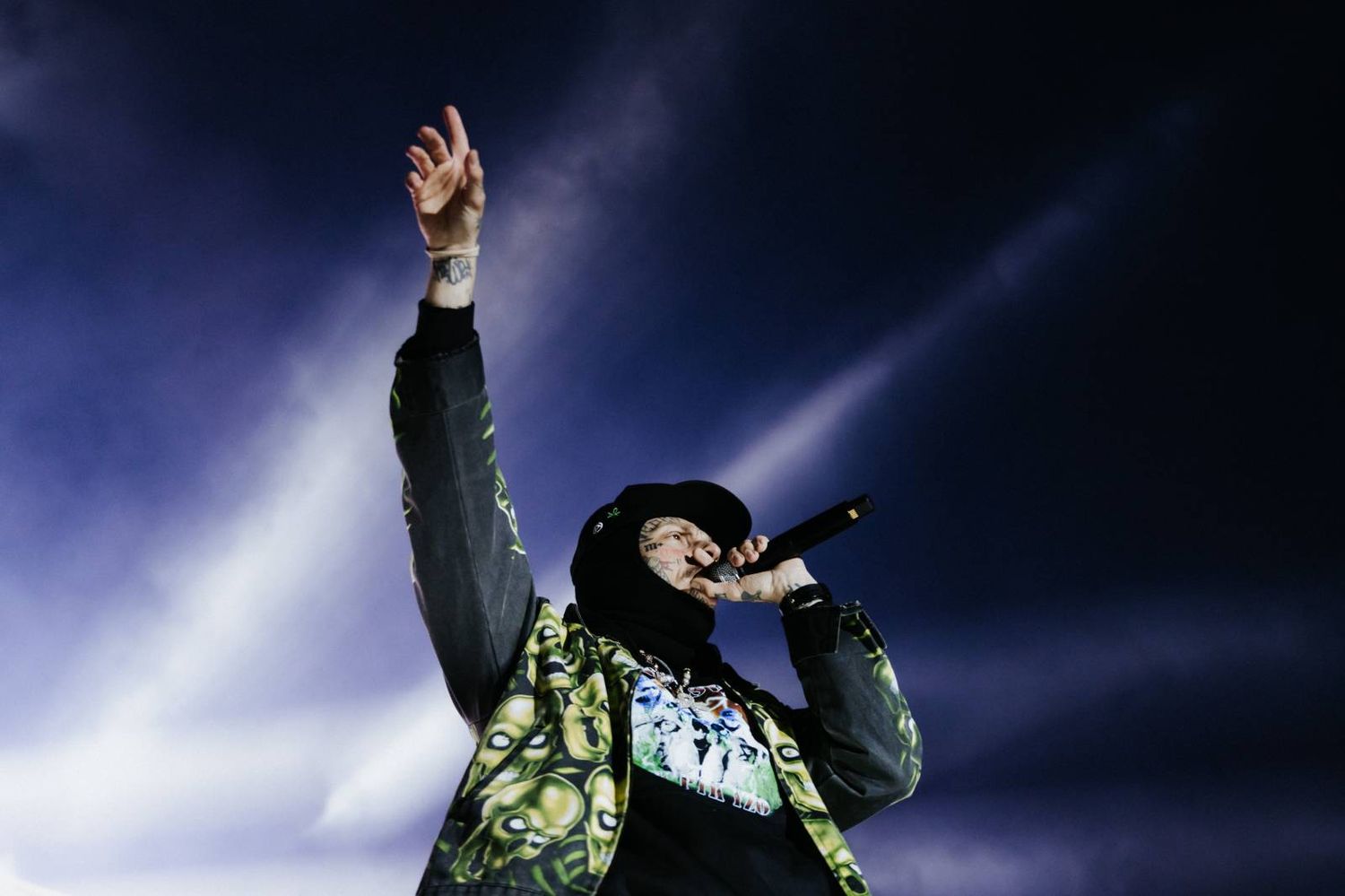 A performer in a dark jacket raises their arms on stage with dramatic lighting and smoke effects in the background.