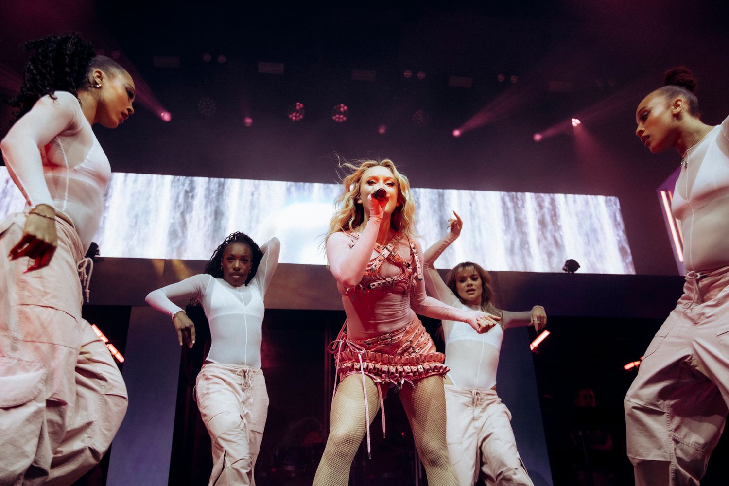 Dancers in white costumes perform an energetic dance routine on a dark stage with dramatic lighting effects.