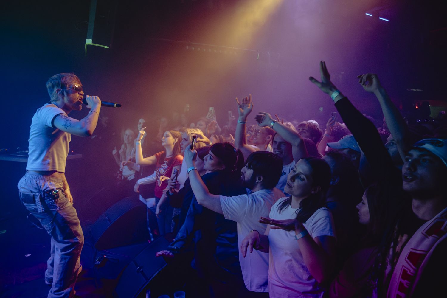 A performer on stage energetically engages with an enthusiastic crowd during a concert under dramatic purple lighting.