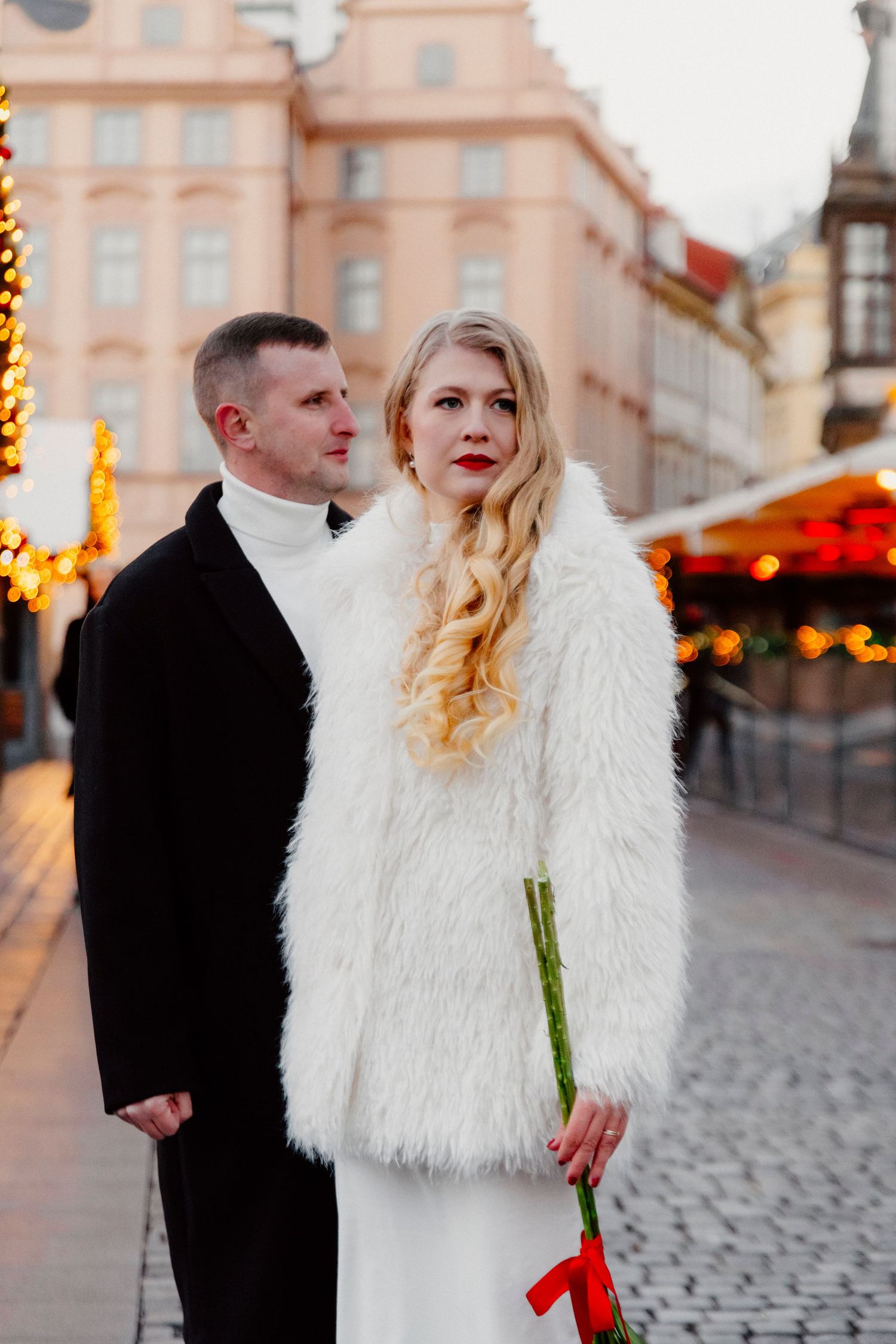Couple in winter wedding attire posing near holiday lights in European city square.