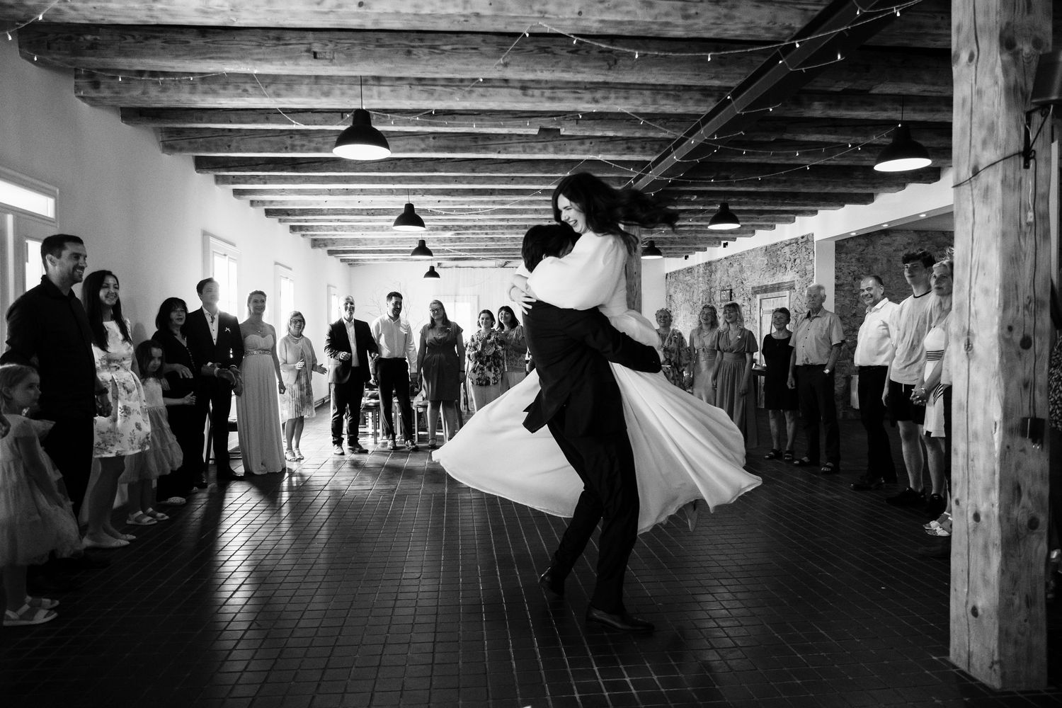 A panoramic black and white shot of people dancing in an indoor reception venue with wooden beams.
