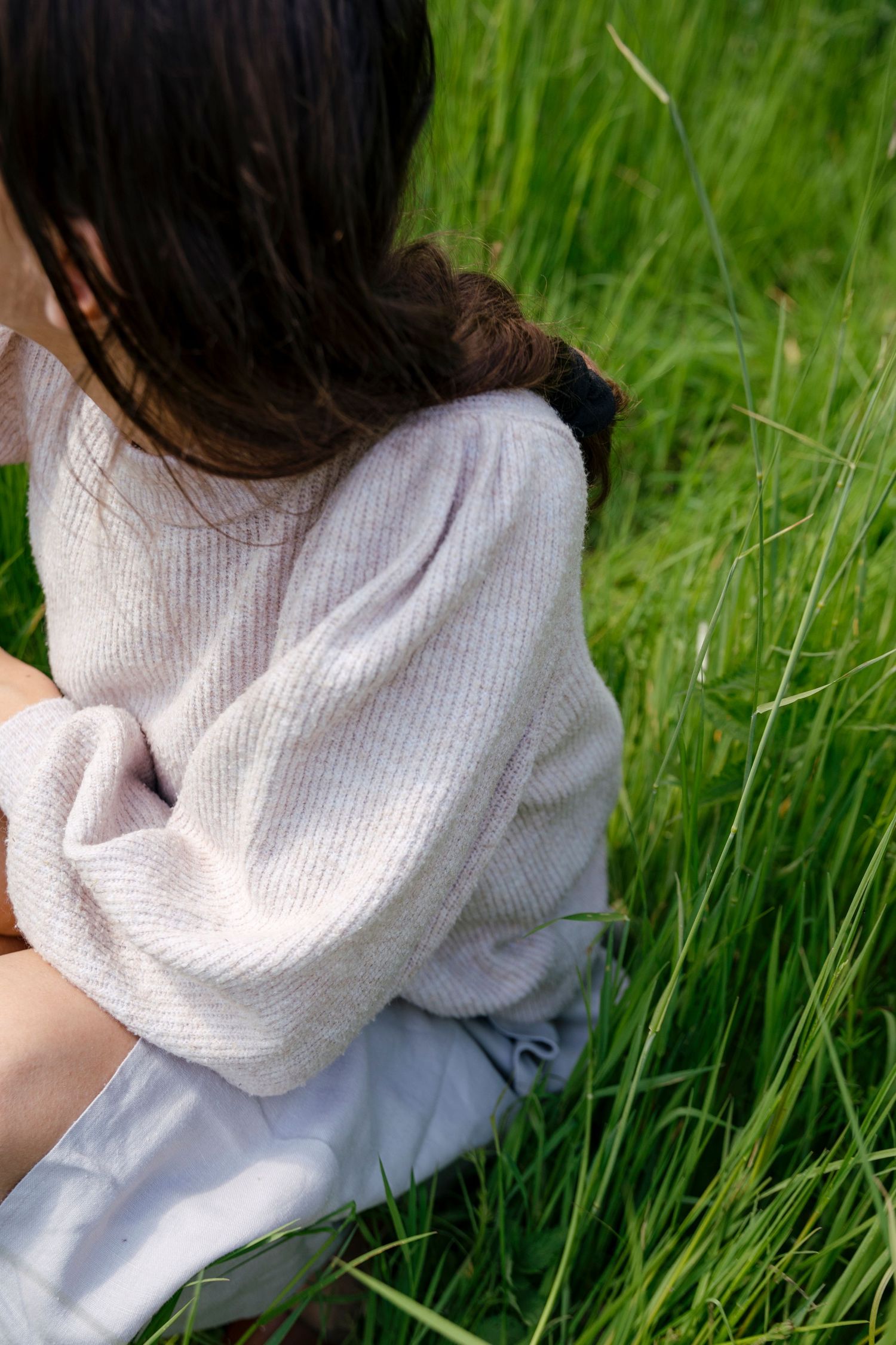 Person in a cozy white sweater sitting in tall grass on a sunny day.