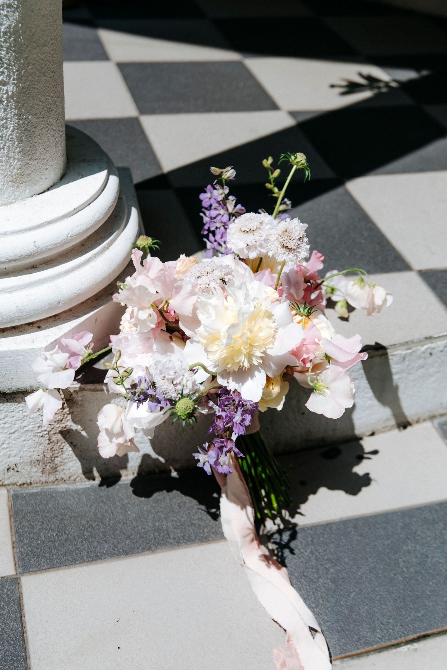 A delicate bridal bouquet with pink and white peonies and purple lilacs rests on a checkered floor in sunlight.