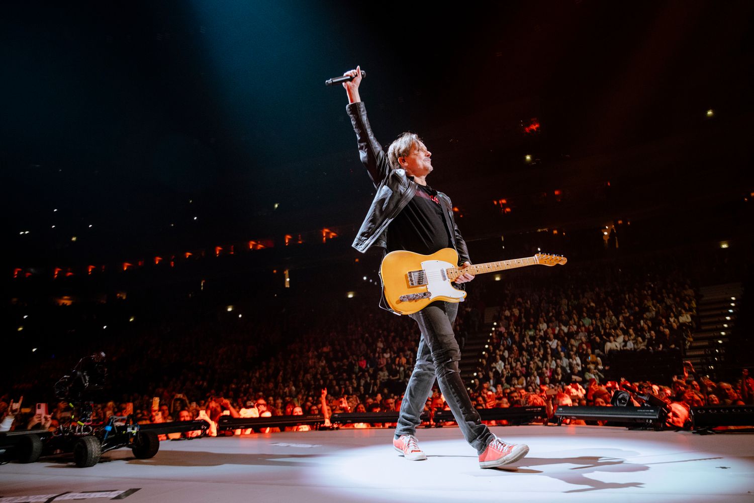 A musician performs on stage in a dark arena with red lighting and a guitar during a live concert performance.