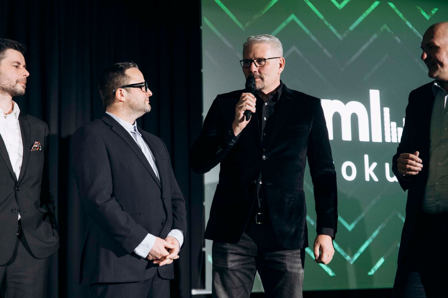 Business professionals in suits converse during a networking event against a green geometric patterned backdrop.