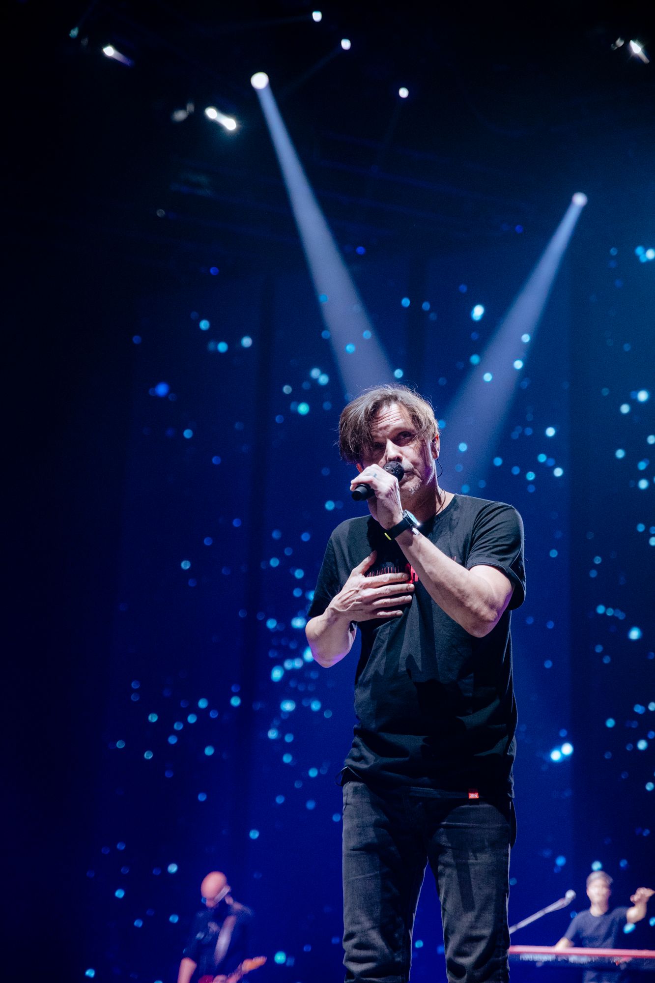 Performer in dark clothing sings passionately on stage with dramatic blue spotlights and sparkles in the background.