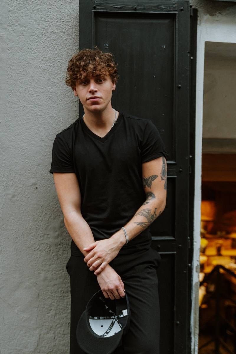 Figure in all black clothing stands against textured wall beside black door frame.