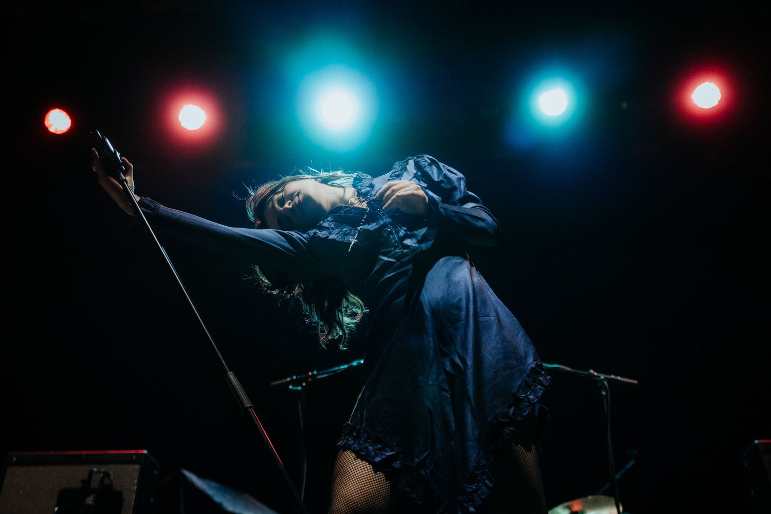 A performer on stage leans back dramatically while singing into a microphone under red and blue stage lights.