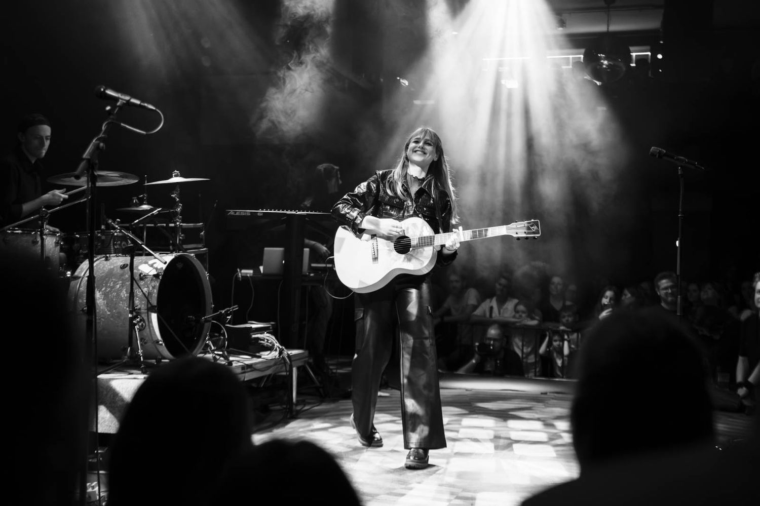 A musician performs with an acoustic guitar on a dimly lit stage with dramatic spotlights and a drum kit visible in the background.