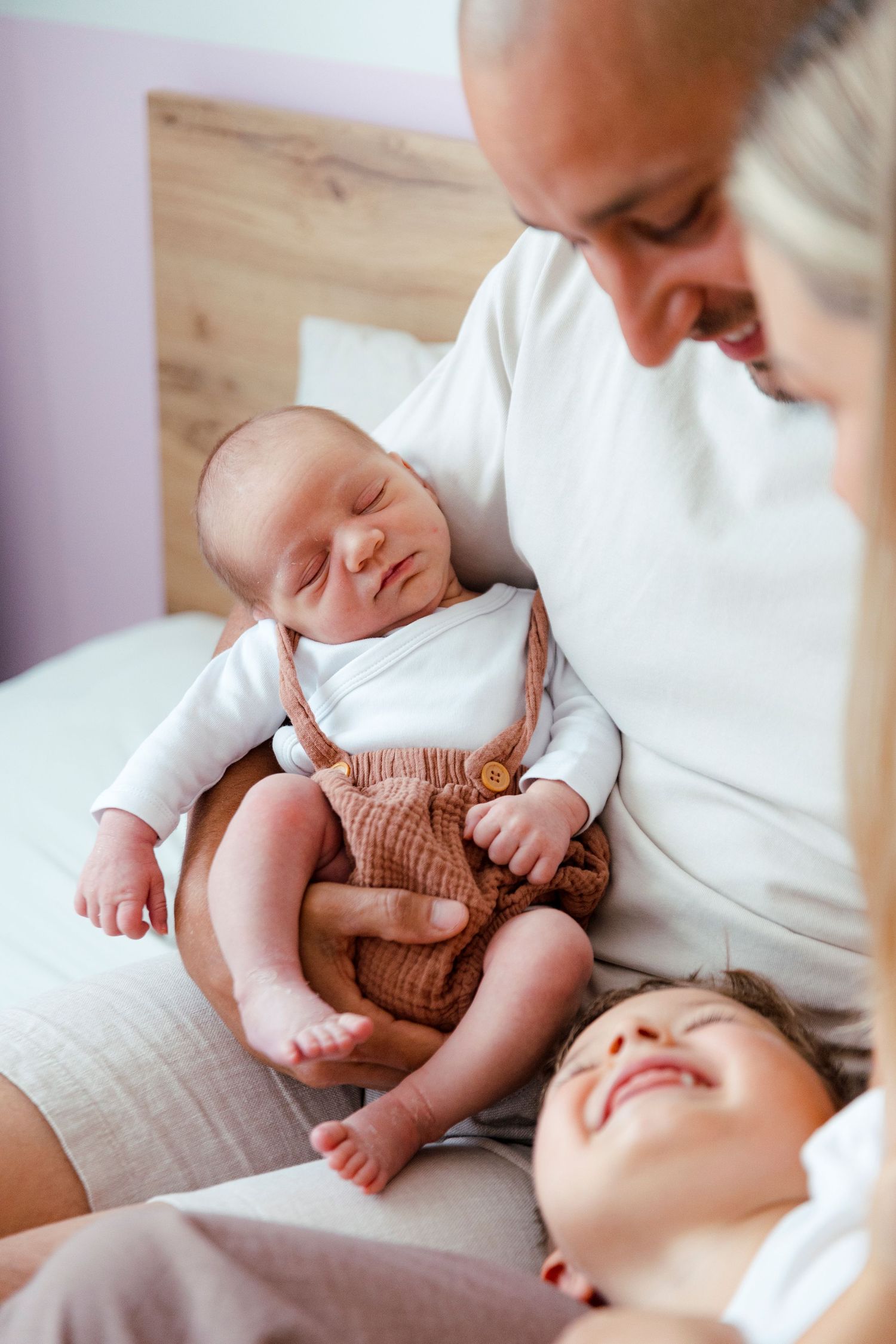 Parents sitting on bed lovingly gazing at their sleeping newborn baby in brown overalls with toddler nearby.