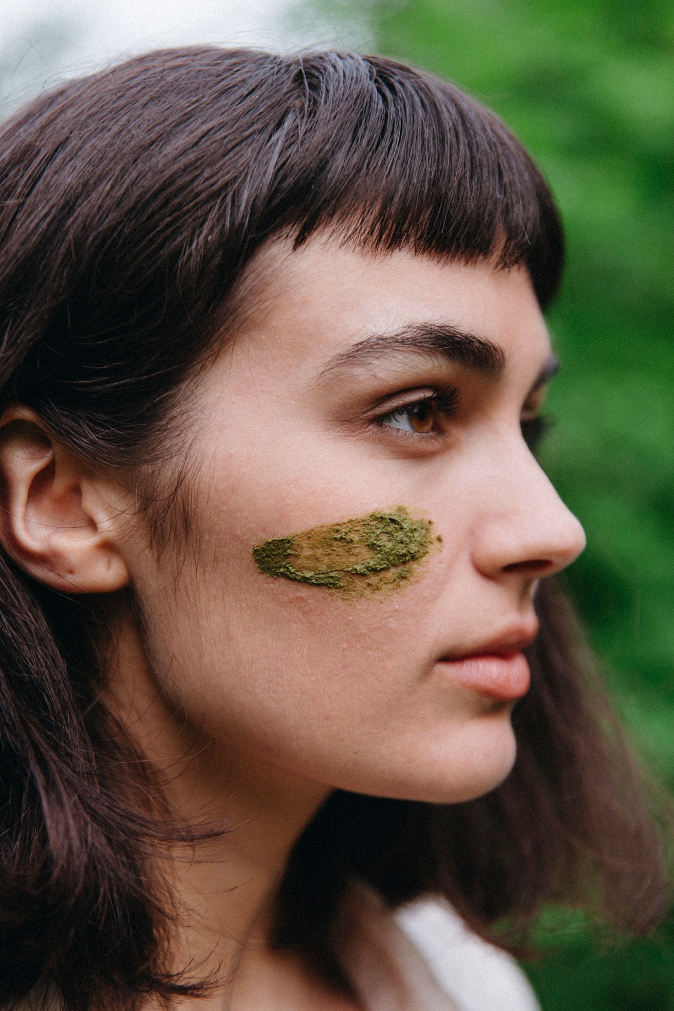 Artistic portrait series showing green natural material applied to face against blurred background.