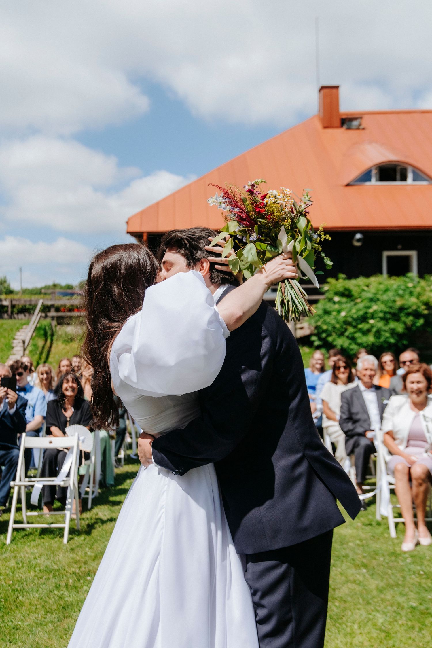 A couple shares their first dance surrounded by guests on a sunny lawn with a terracotta-roofed building in the background.