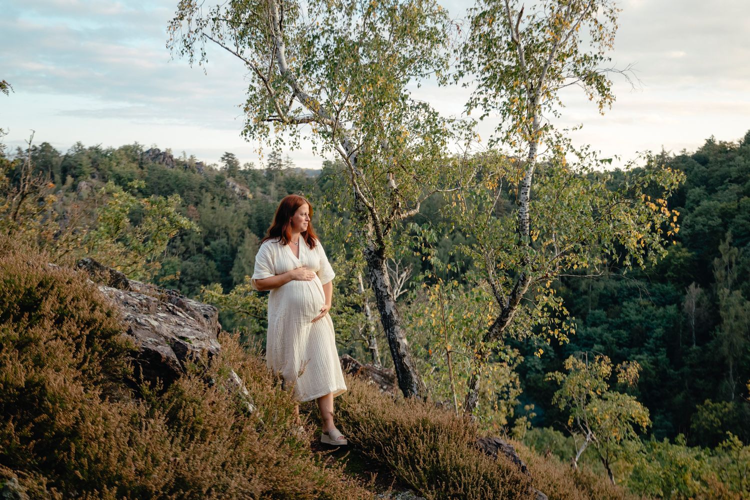 Pregnant woman in flowing dress overlooking valley from mountain viewpoint.