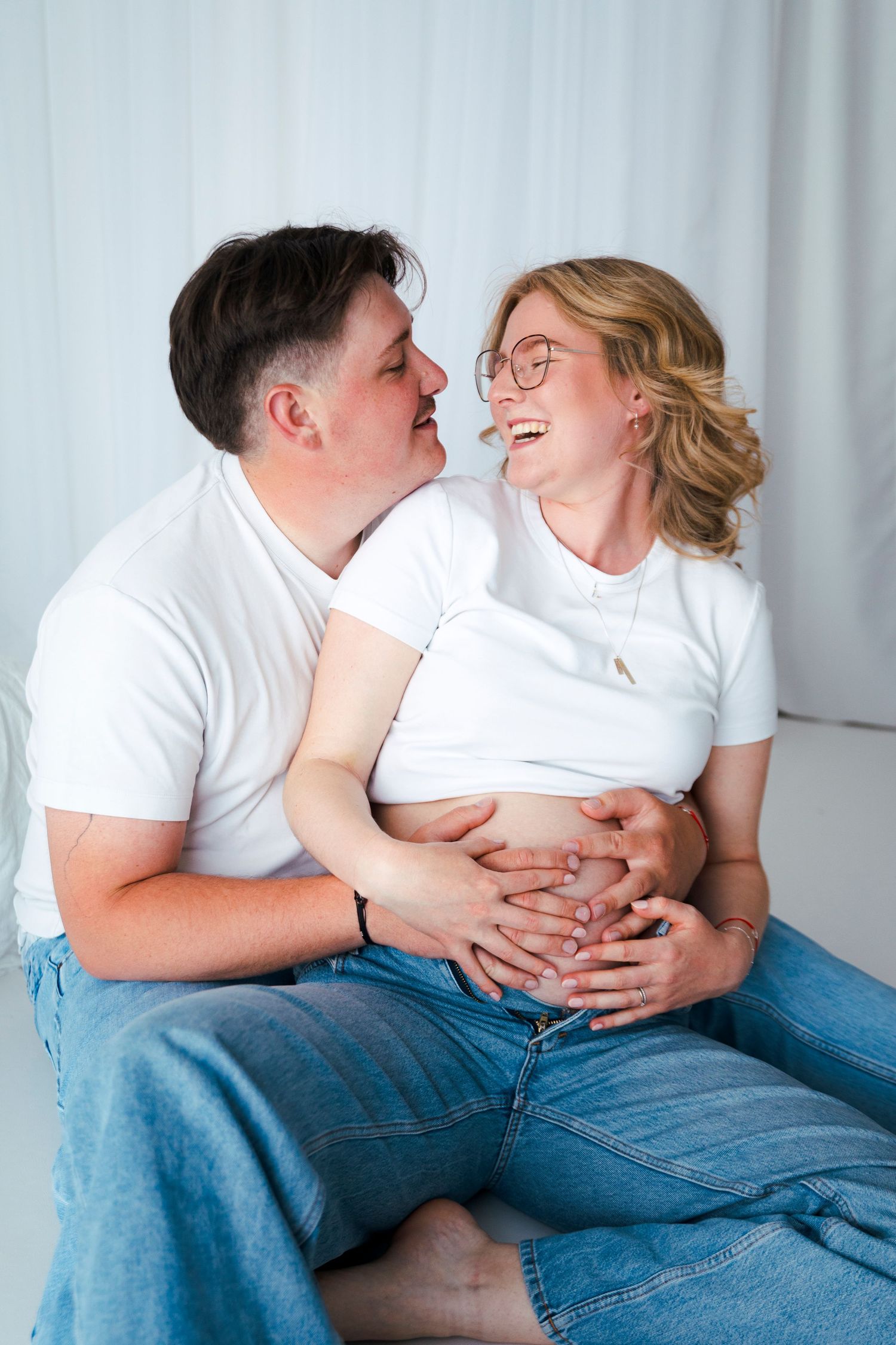 Joyful maternity photo of couple laughing together on white backdrop.