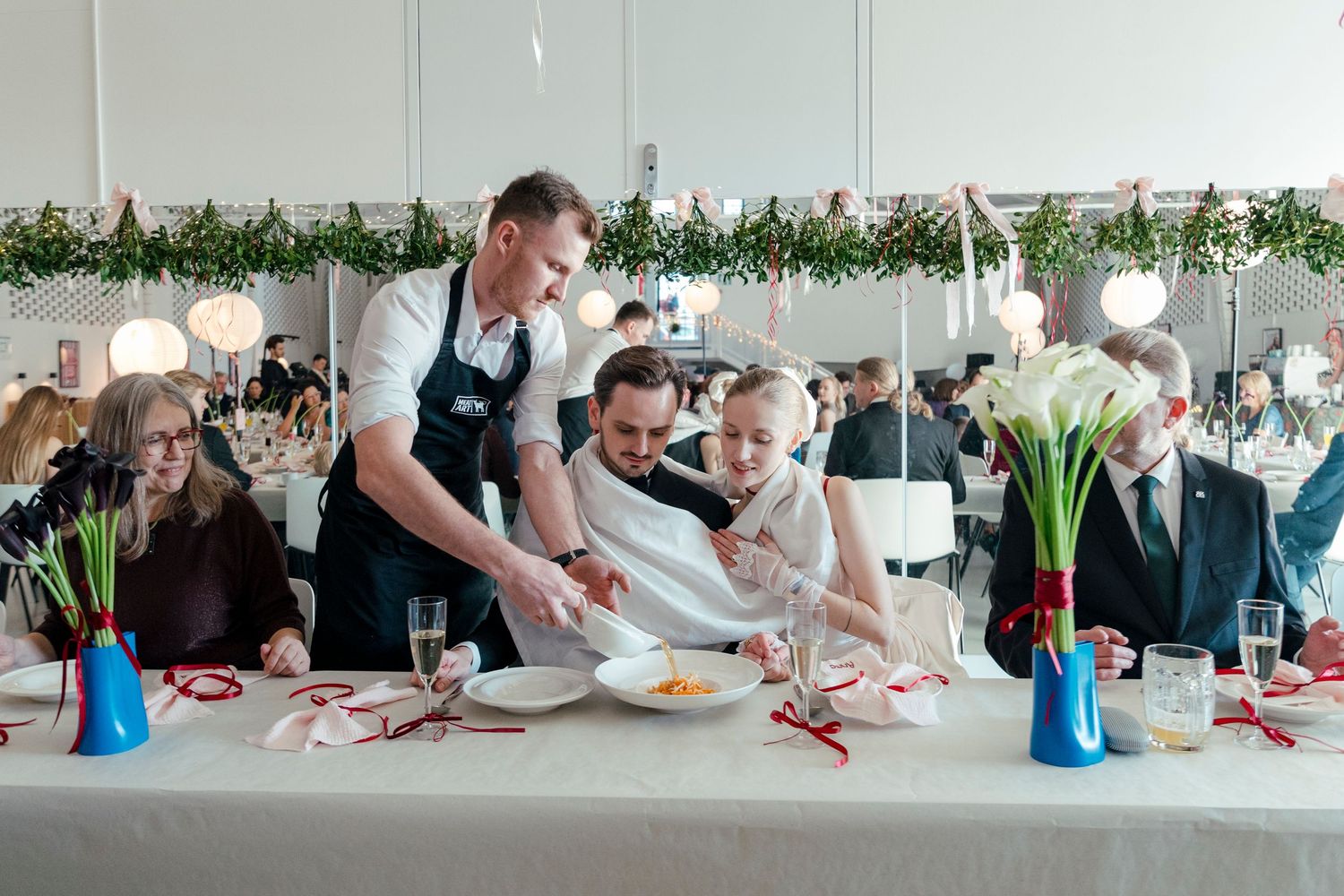 Guests at a wedding reception gather around a decorated table with blue vases and floral arrangements while being served.