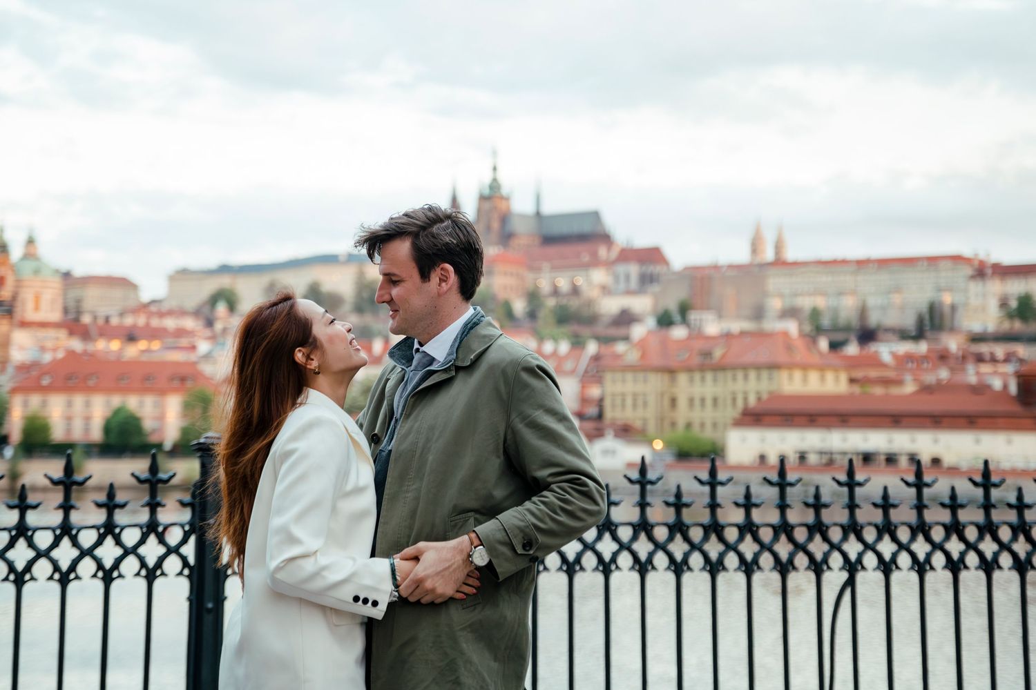 A romantic couple stands along a historic city overlook with European architecture in the background.