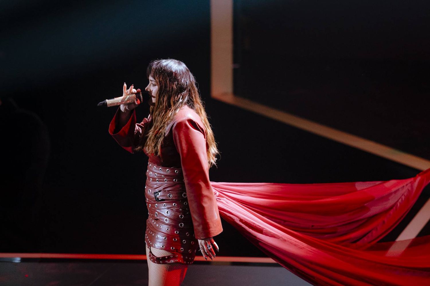 A performer in a flowing red dress and cape sings passionately on a dimly lit stage.