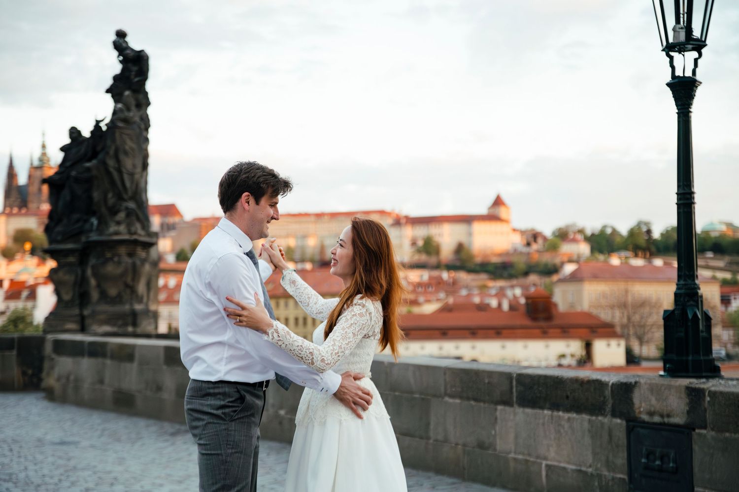 Romantic couple embraces on Charles Bridge in Prague with historic architecture and cityscape in background.