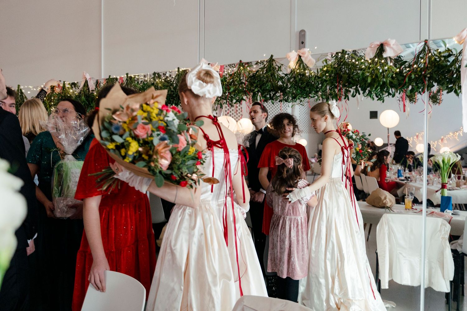 Wedding guests in formal attire gather and socialize at an indoor reception decorated with greenery and red accents.
