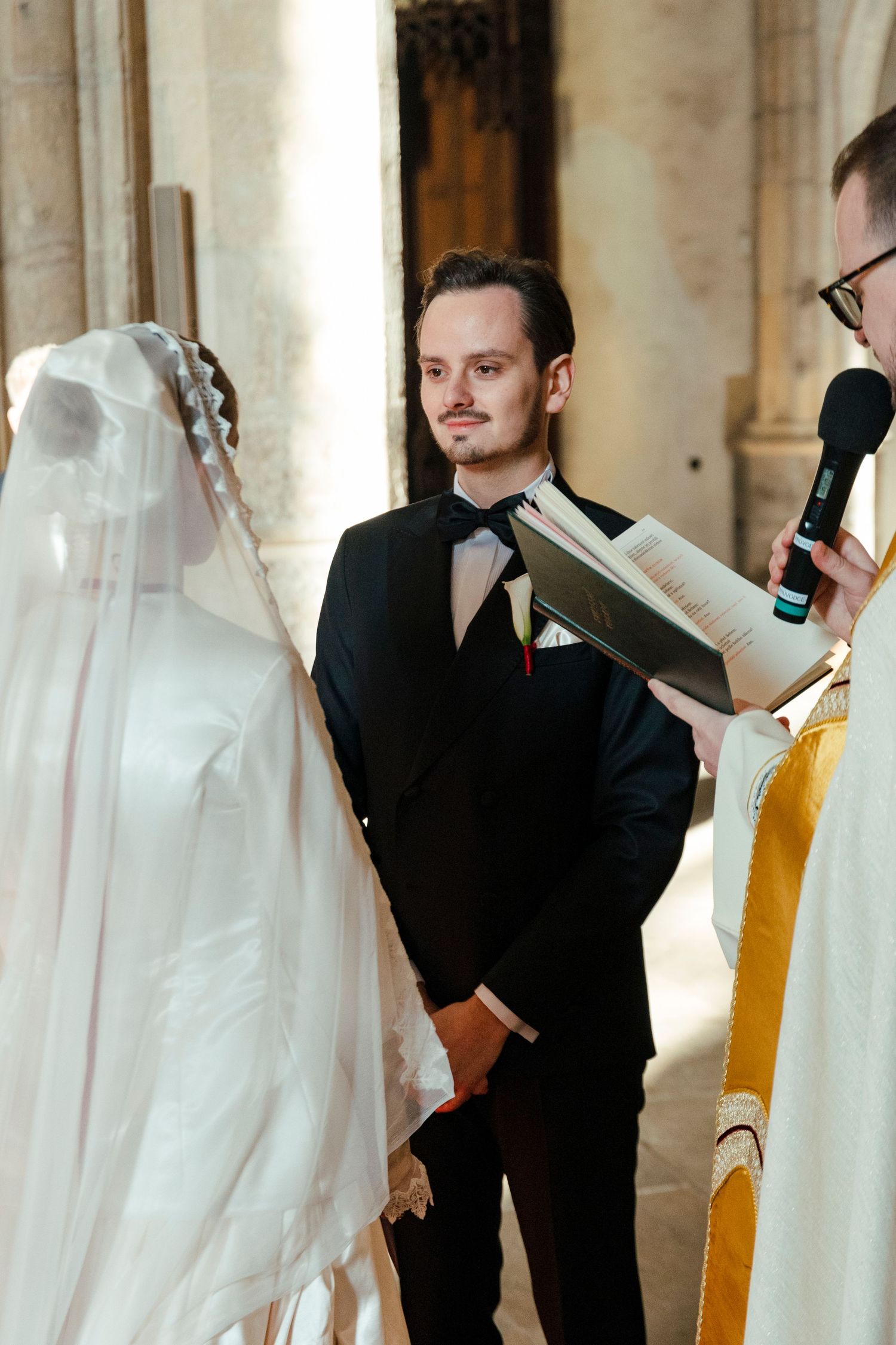 Wedding party members in formal attire checking their phones while standing in an elegant columned hallway.